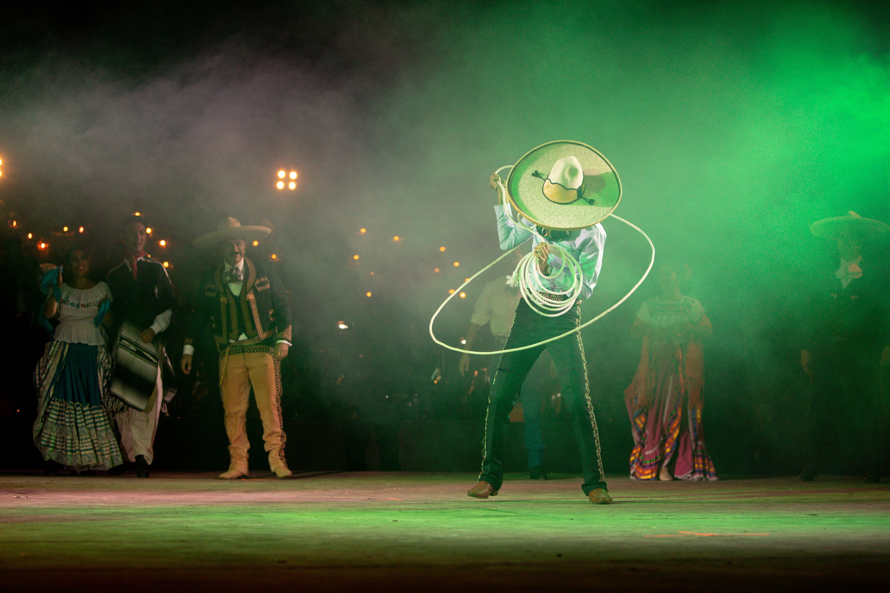 Charro con reata rodeado de elenco bajo luces verdes y humo escénico